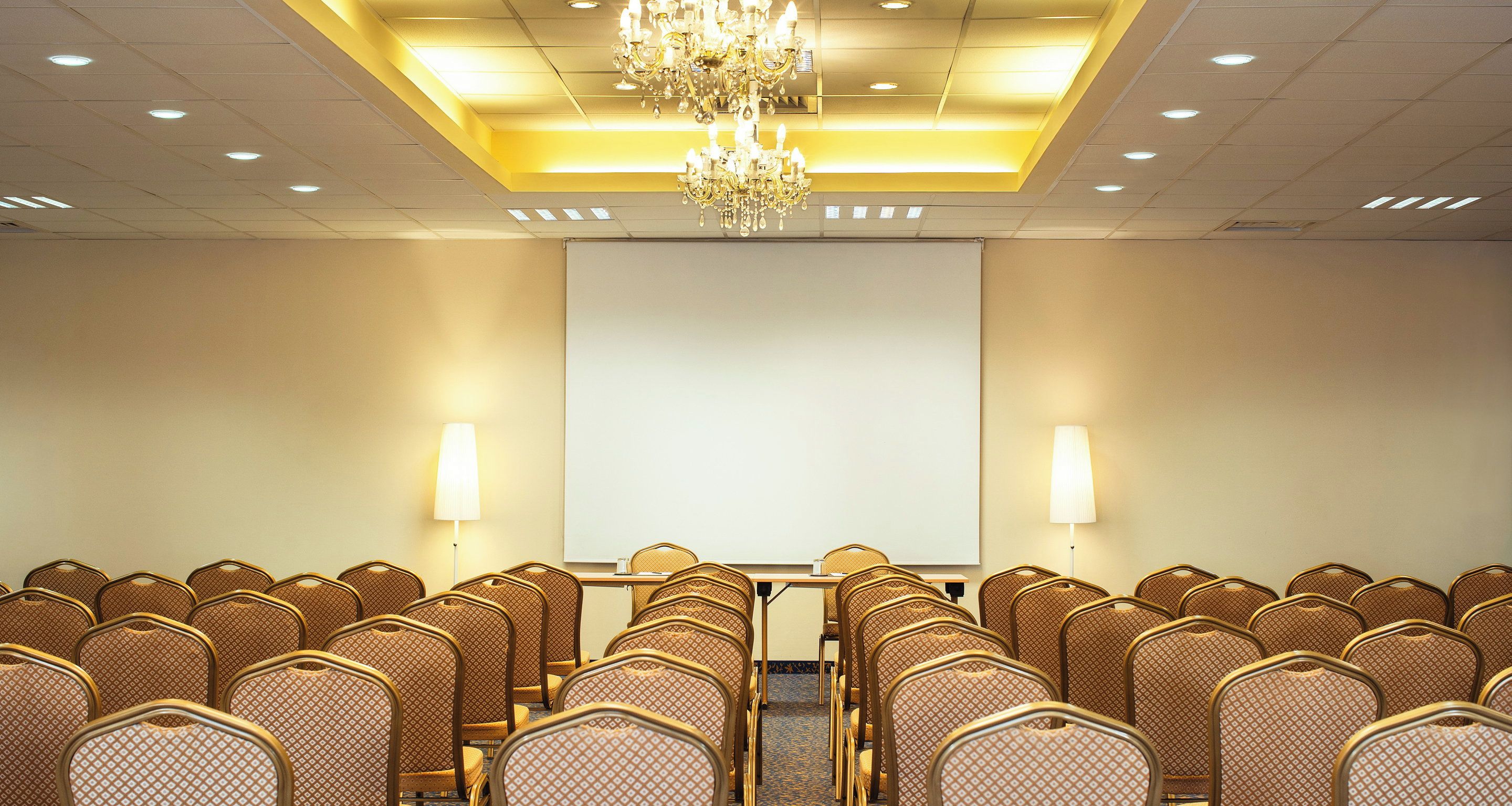 Un salón de conferencias con filas de sillas, una pantalla de proyector, una mesa con una computadora portátil y una araña.