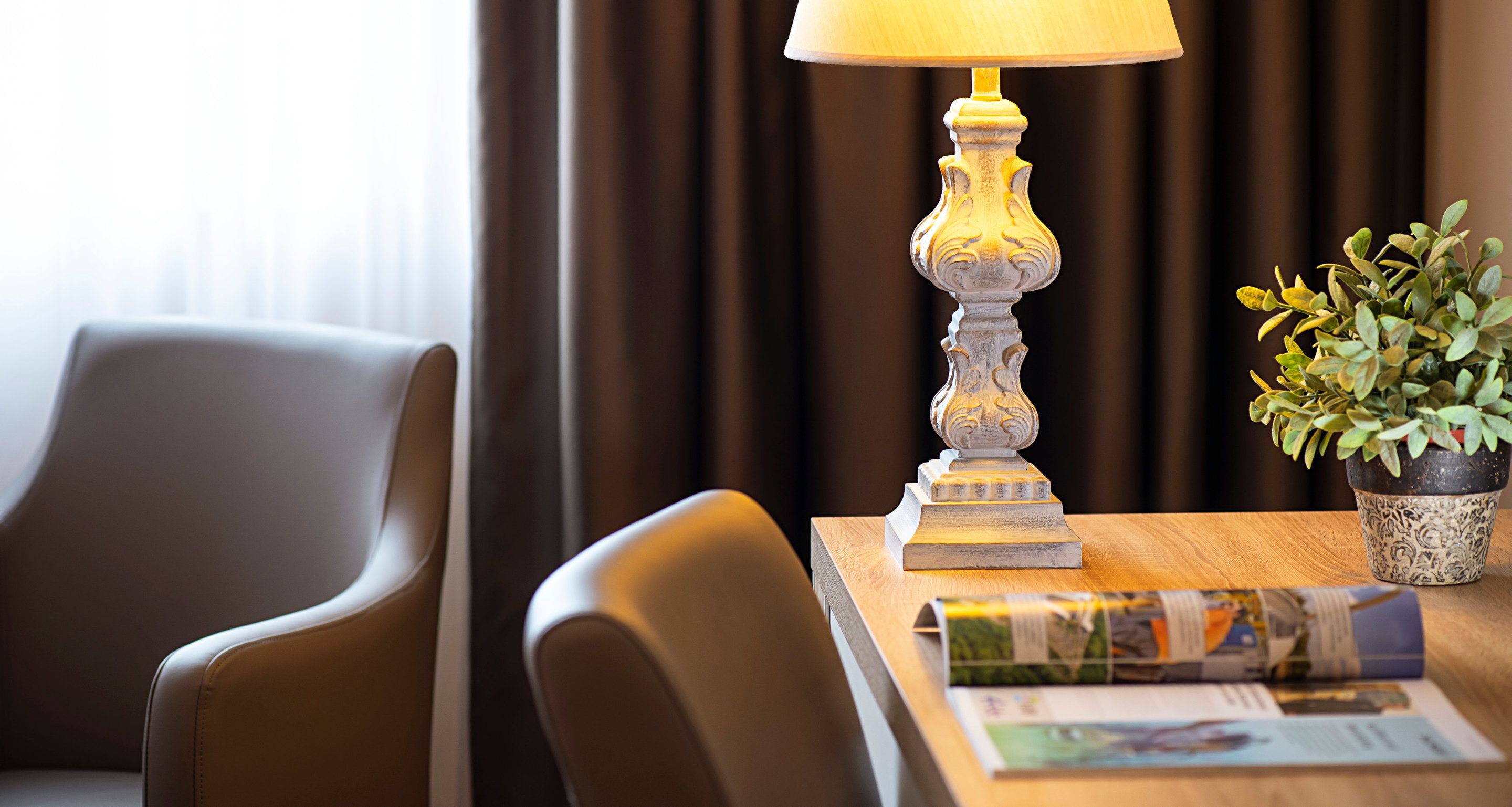 Una habitación de hotel con una mesa de madera, una lámpara, una planta y dos sillas.