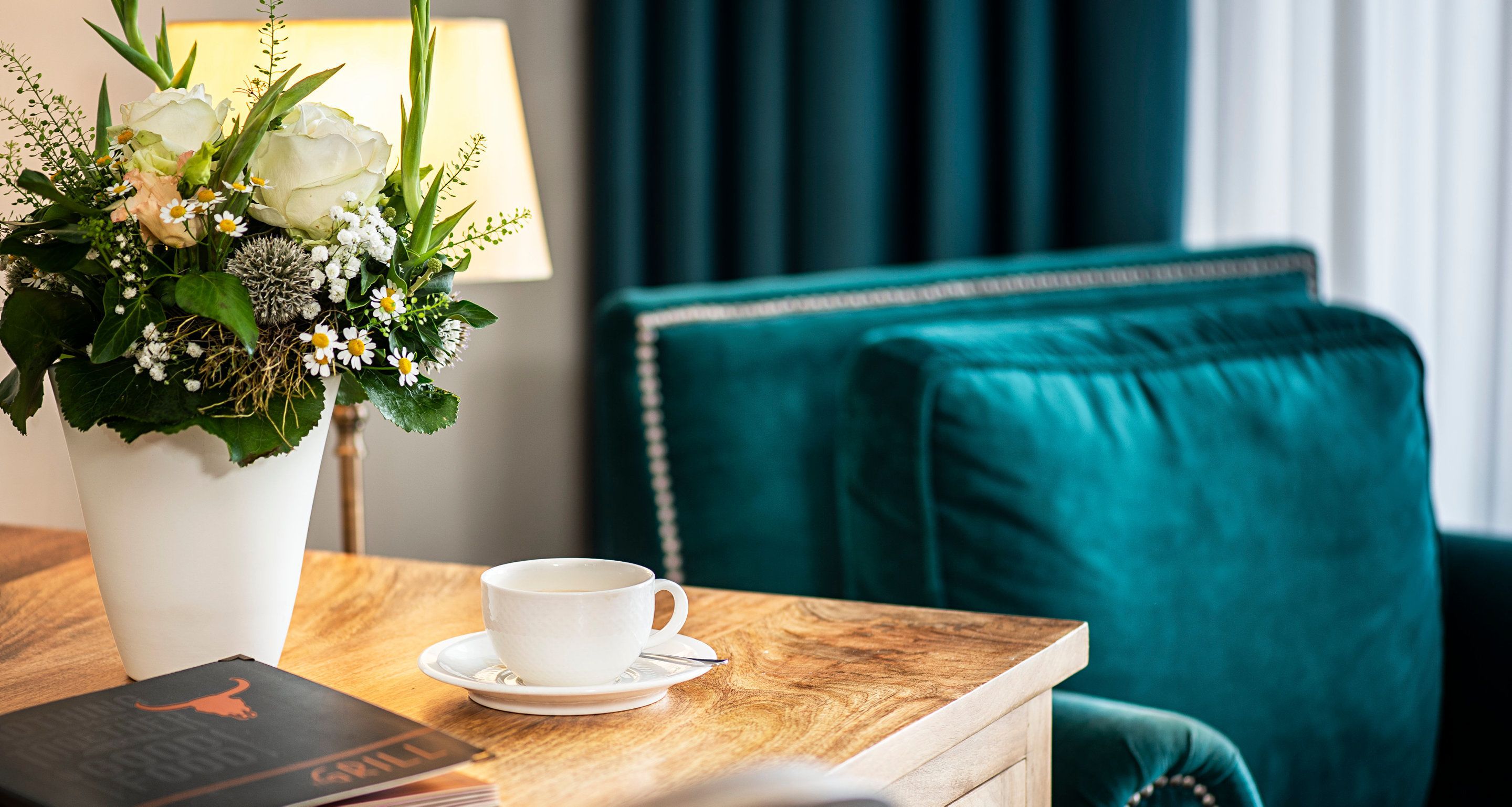 A room with a wooden table, a white cup and saucer, a book, a vase with flowers, and teal upholstered furniture.
