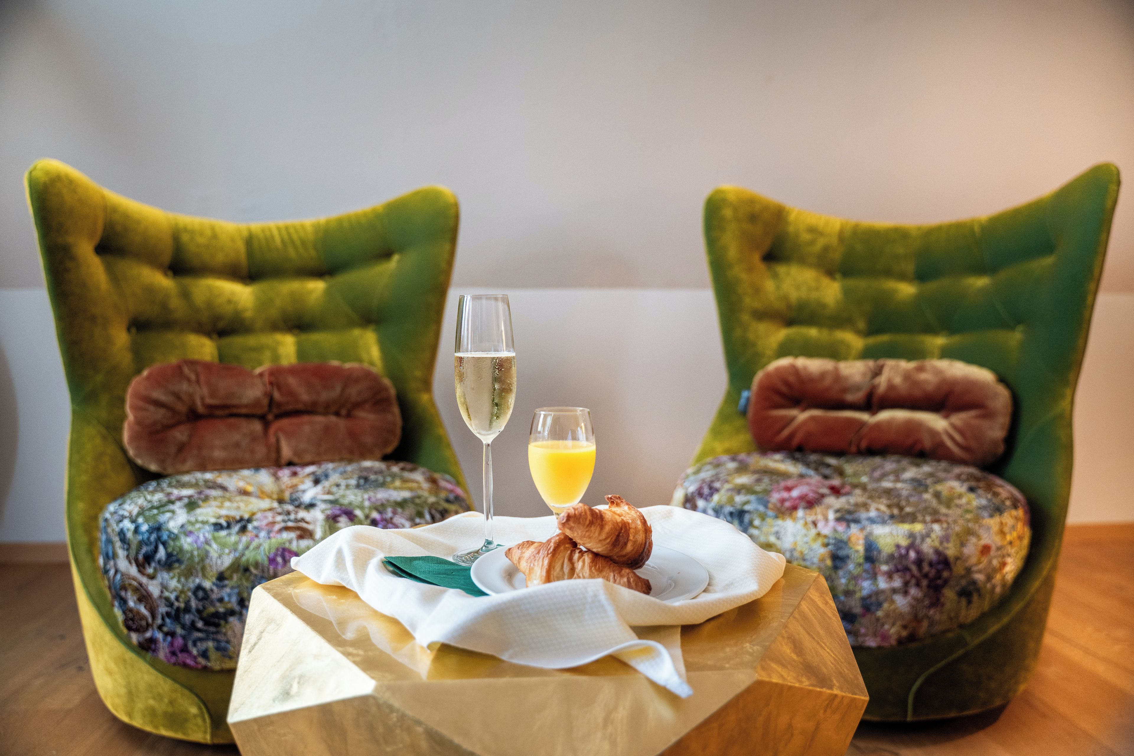Zwei gepolsterte Stühle mit Blumenmuster, ein kleiner Holztisch mit einer Serviette, ein Glas Orangensaft, ein Glas Champagner und ein Croissant.