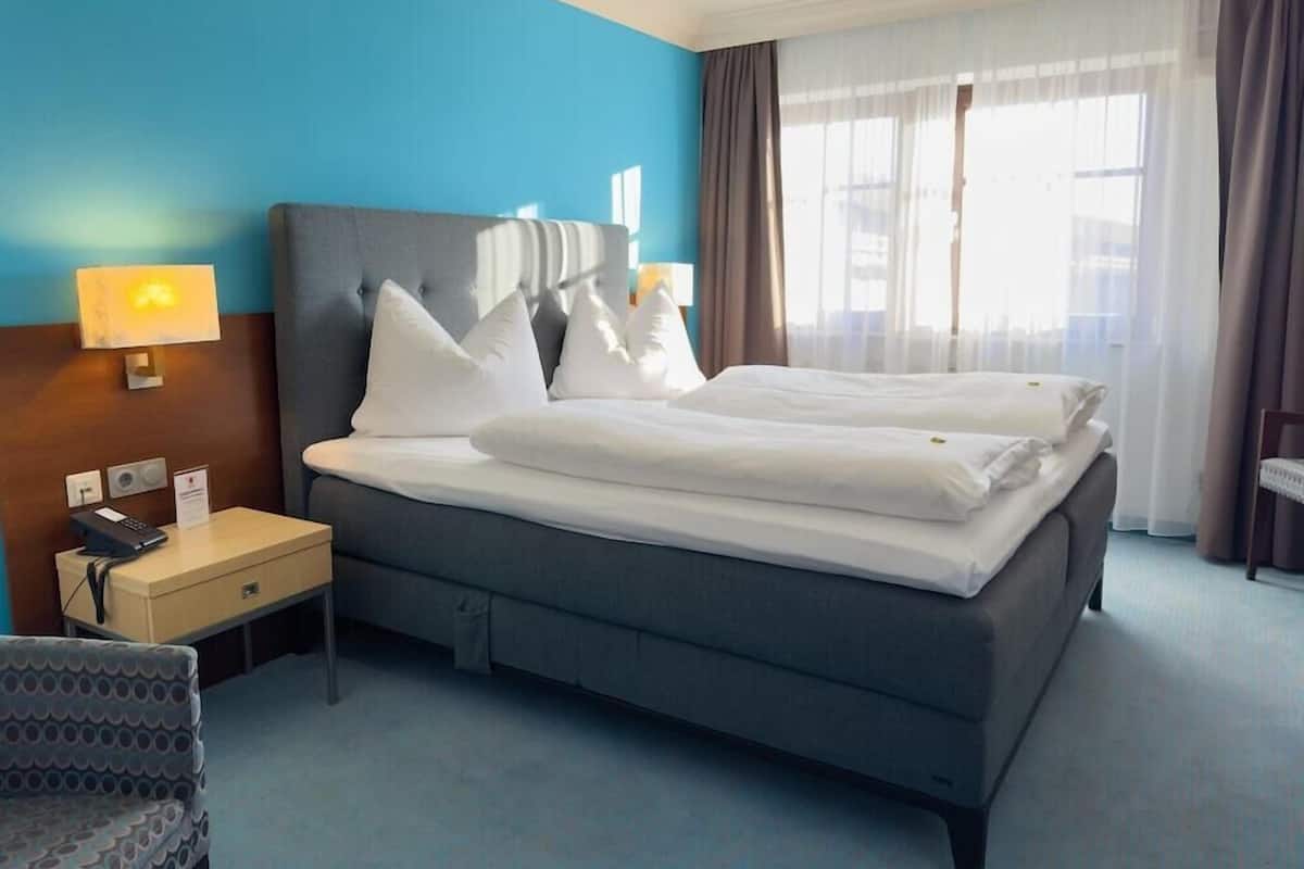 Habitación de hotel con una cama grande, una mesita de noche con lámpara, una silla y vistas a la ciudad por la ventana.