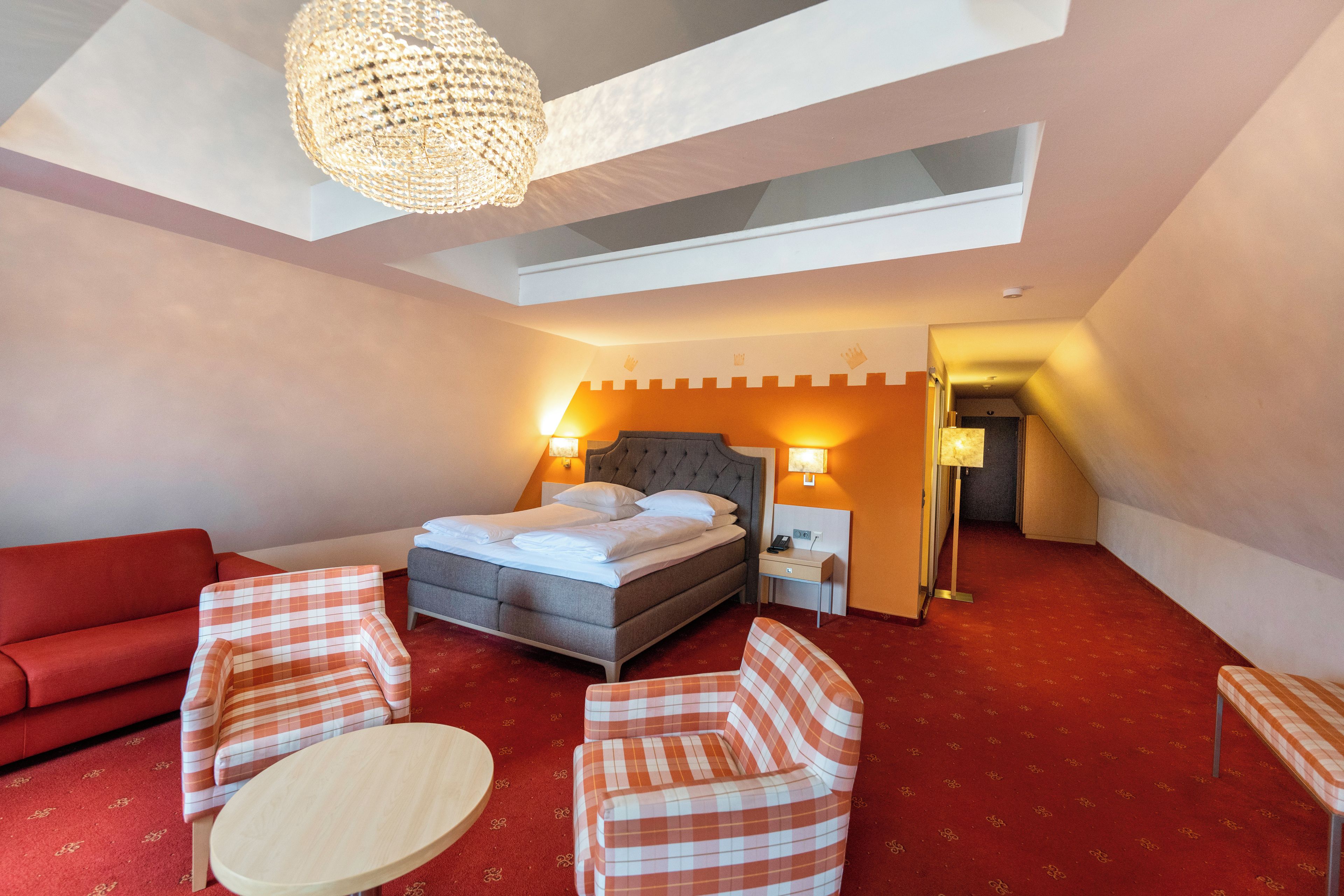 Une chambre d’hôtel avec un lit, deux tables de chevet, un canapé rouge, deux fauteuils à motifs écossais, une table ronde et un lustre.