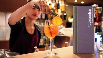 Un cantinero preparando una bebida con una botella etiquetada 'Aperol' y un vaso con una rodaja de naranja. Un menú titulado 'topinambar' es visible sobre la mesa.