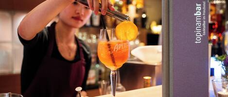 Un barman prépare un cocktail à l’aide d’une bouteille étiquetée « Aperol » et d’un verre dans lequel on a mis une tranche d’orange. On peut voir sur la table un menu intitulé « topinambar ».