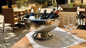 Un espacio de comedor al aire libre con mobiliario de mimbre, un recipiente metálico con botellas de vino y un paraguas verde.