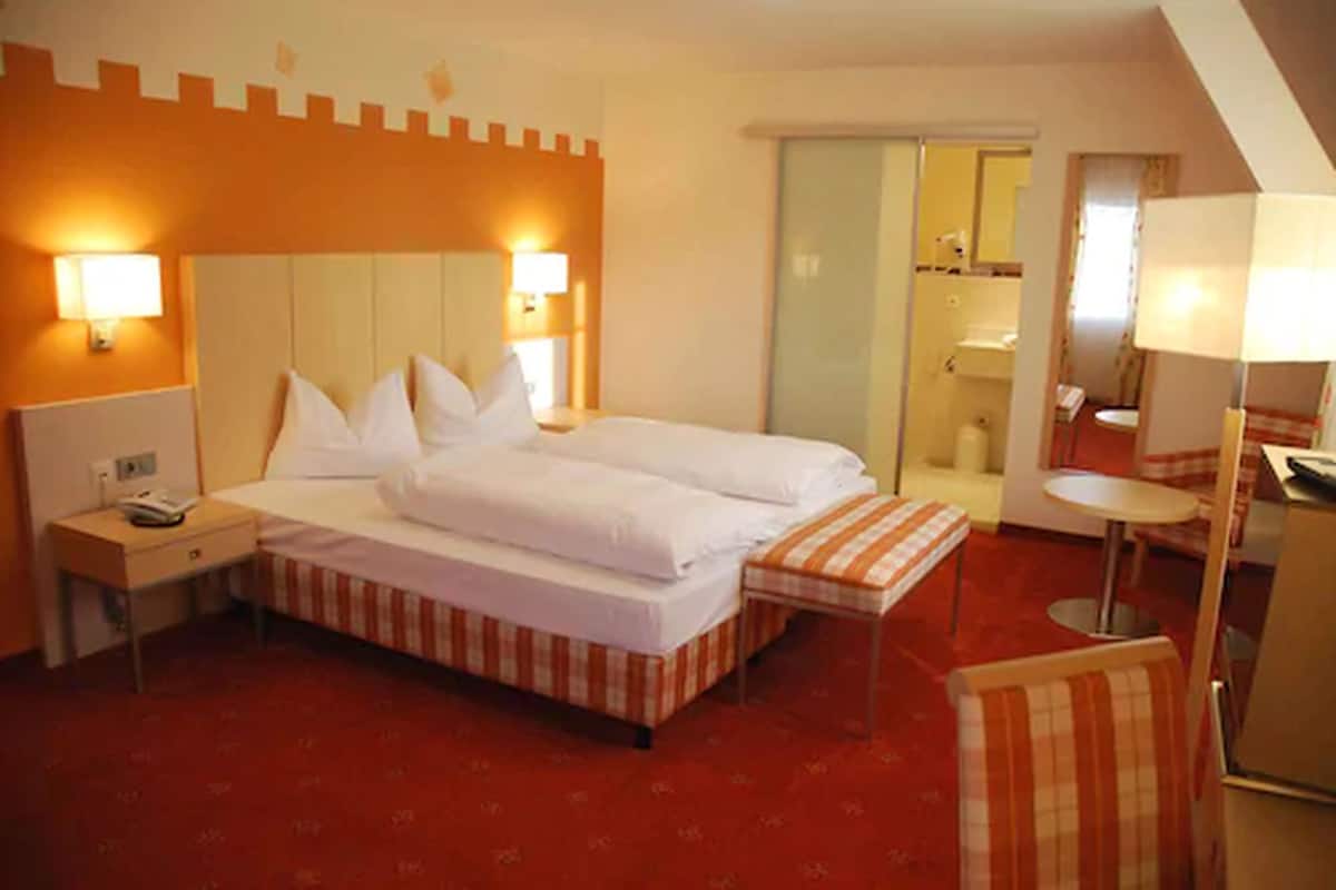 Habitación de hotel con cama, mesitas de noche, una pequeña mesa, una silla, un espejo y un baño visible a través de una puerta.