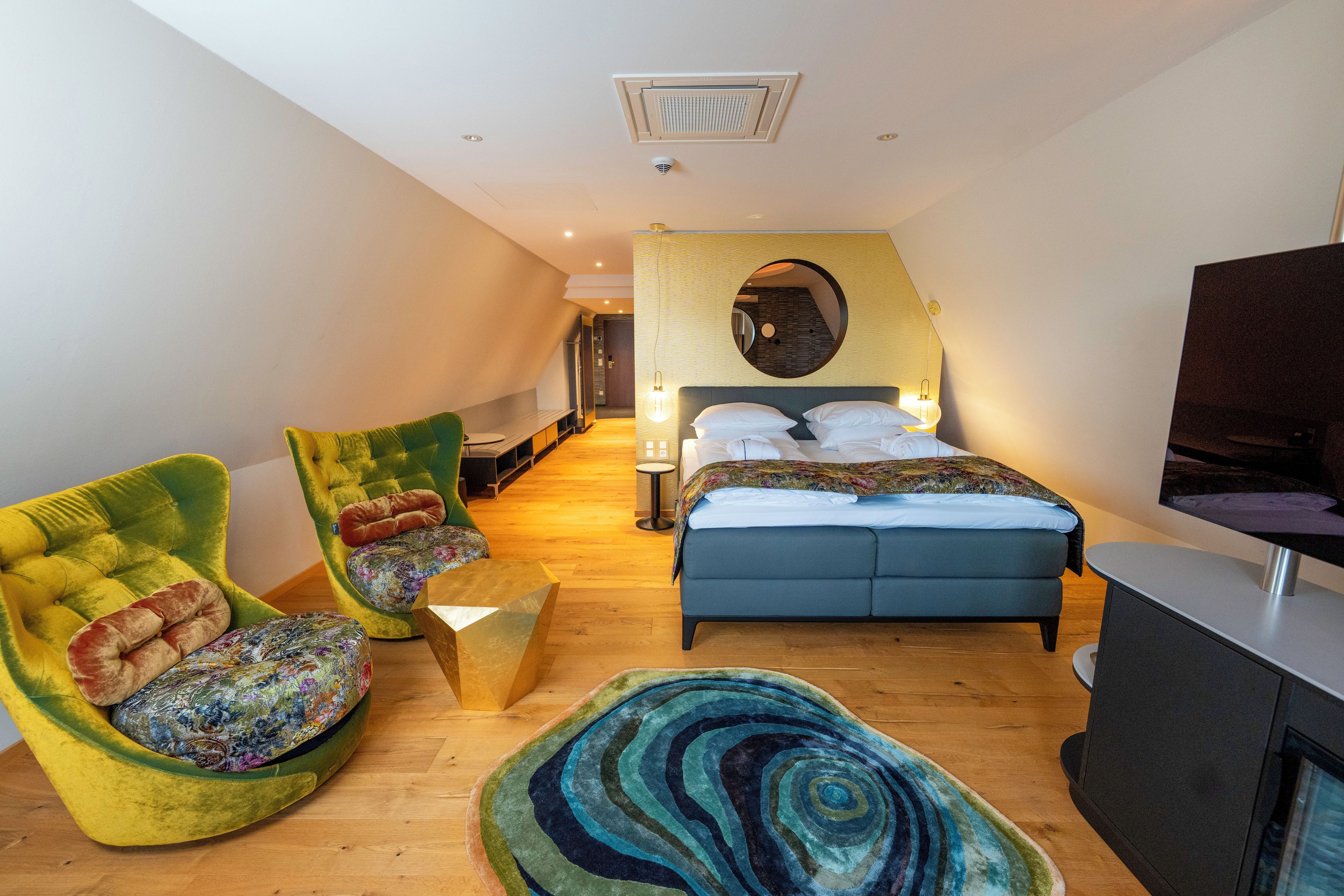 Une chambre d’hôtel moderne avec un lit, un téléviseur, un fauteuil vert, un tapis rond et un plancher en bois.