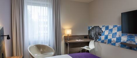 A hotel room with a bed, a desk, a chair, a lamp, and a television.