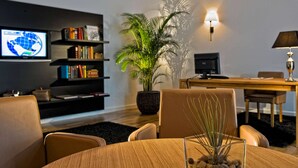 A modern living room with a round wooden table, leather chairs, a bookshelf, a television, a desk, and a potted plant. - ibis Styles Rosenheim (Rosenheim)