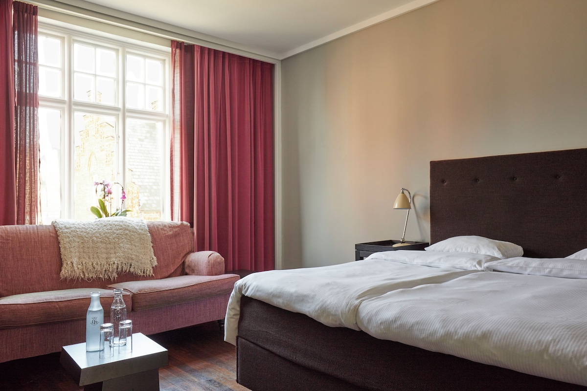 A hotel room with a bed, a sofa, a nightstand, and a window with curtains.