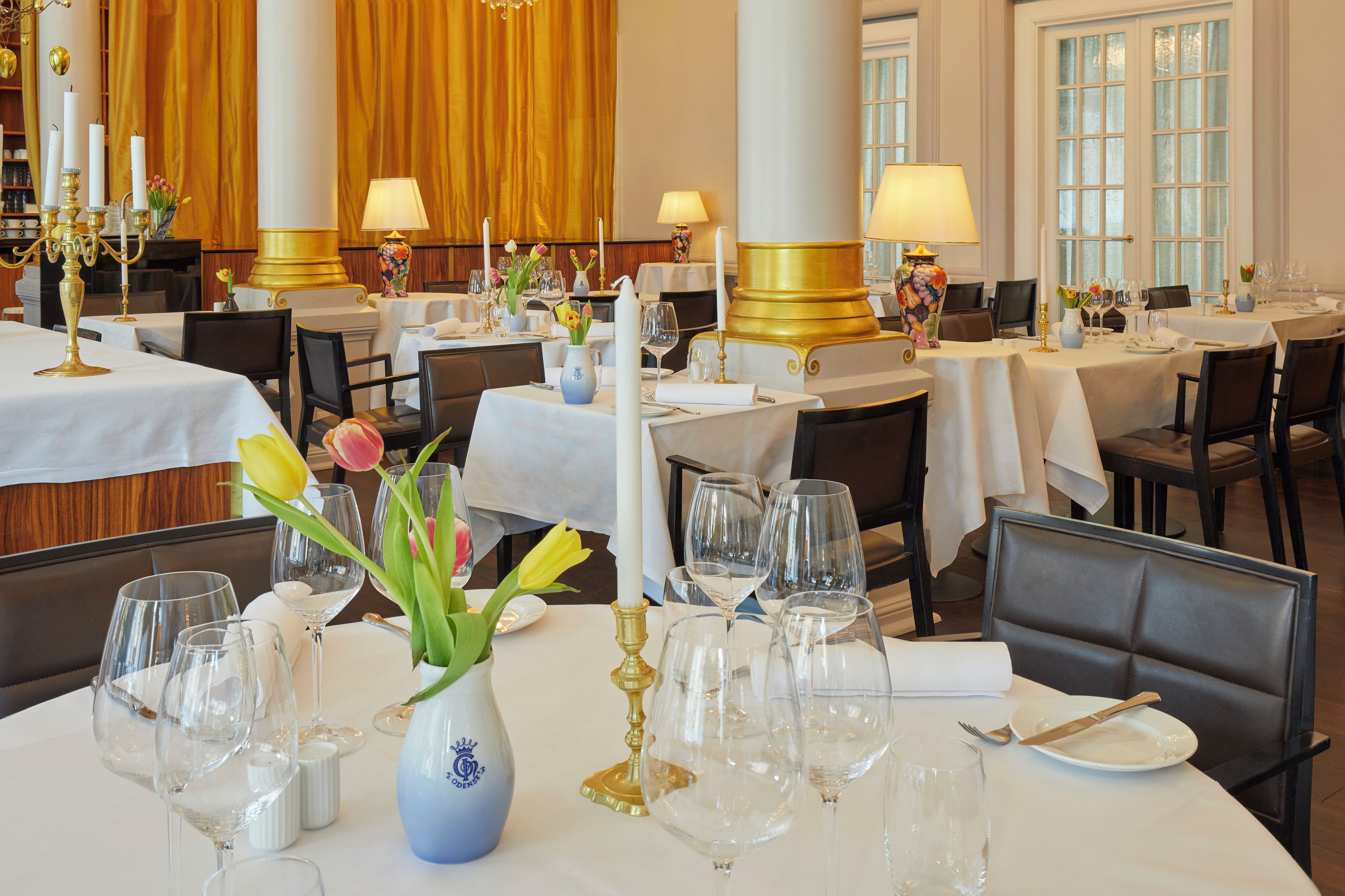 Un comedor formal con mesas dispuestas para el servicio, con manteles blancos, candelabros dorados y centros de mesa florales.