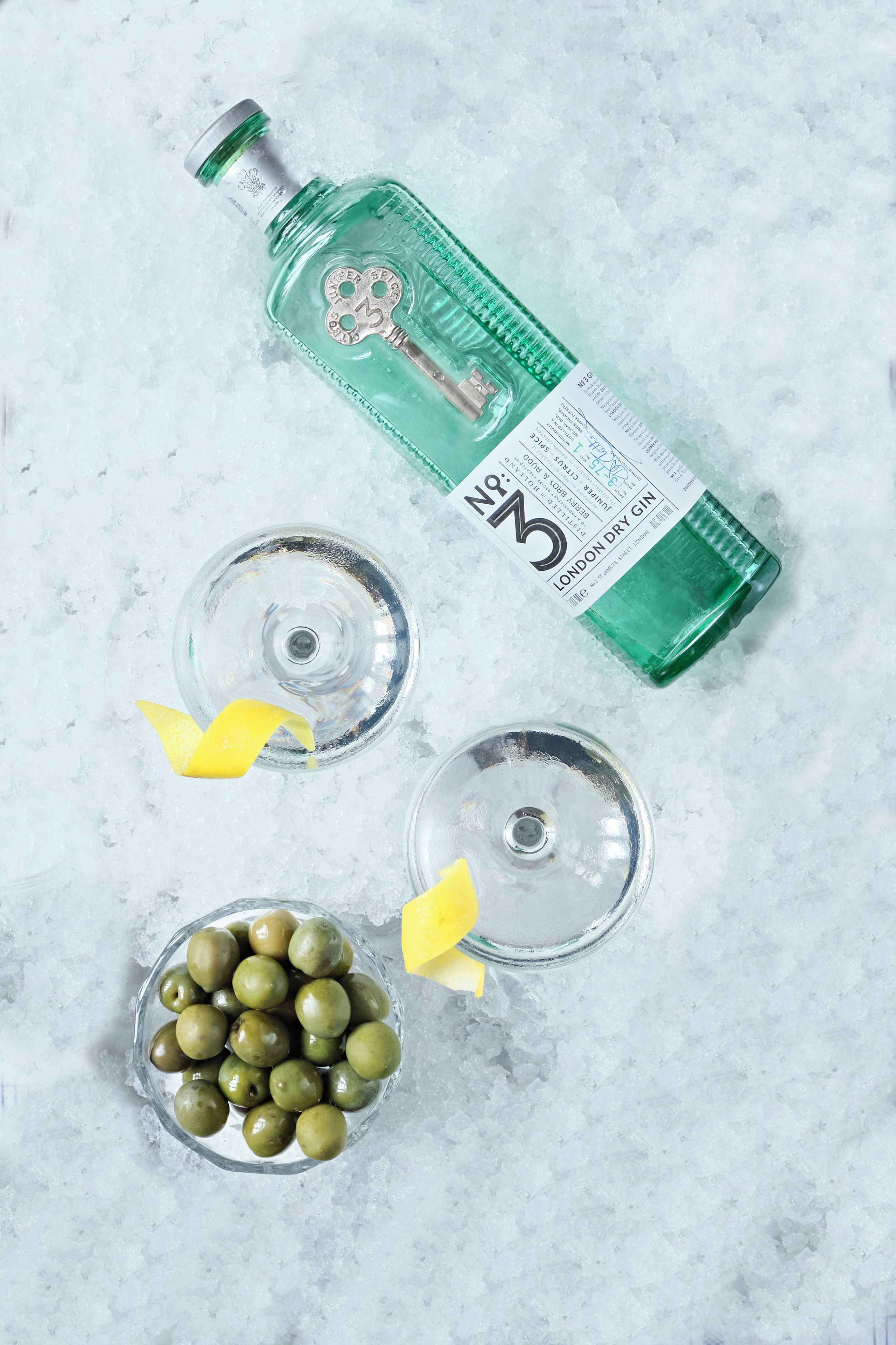 Una botella de ginebra London Dry, dos vasos de vidrio vacíos y un cuenco de aceitunas sobre una cama de hielo.