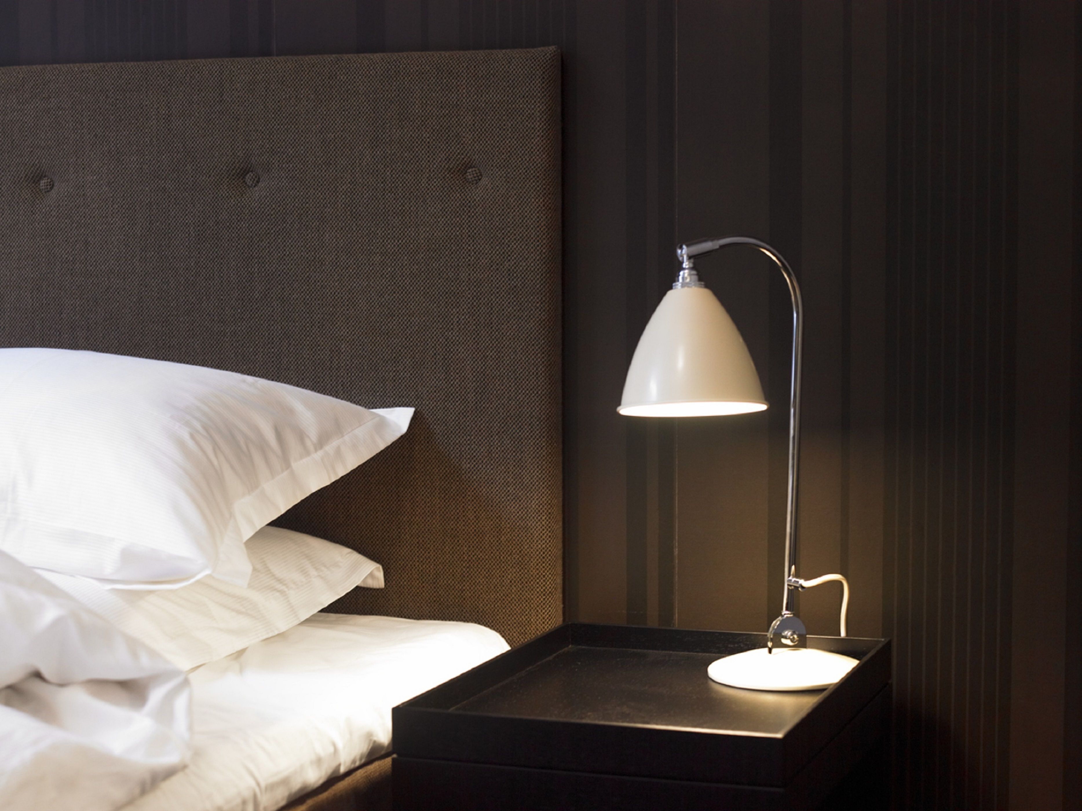 En pænt redt seng med hvide sengetøj og puder, en mørk sengegavl og et natbord med en hvid lampe.