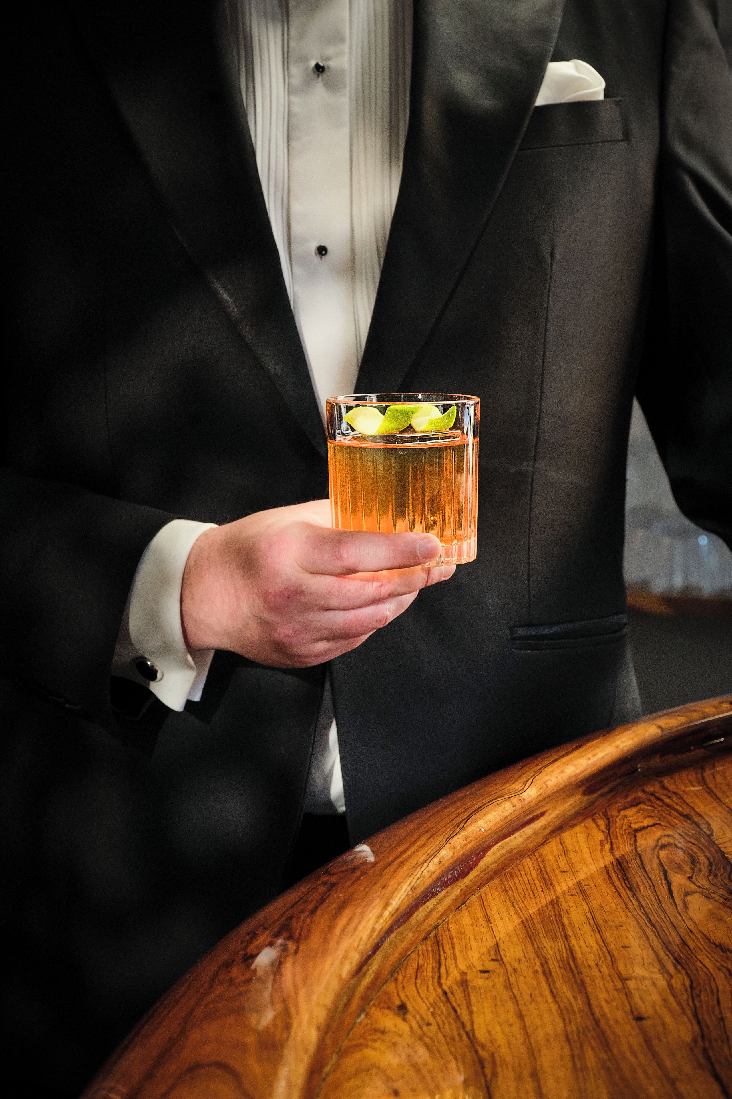 A person in a black suit holding a glass with a drink, standing next to a wooden table.