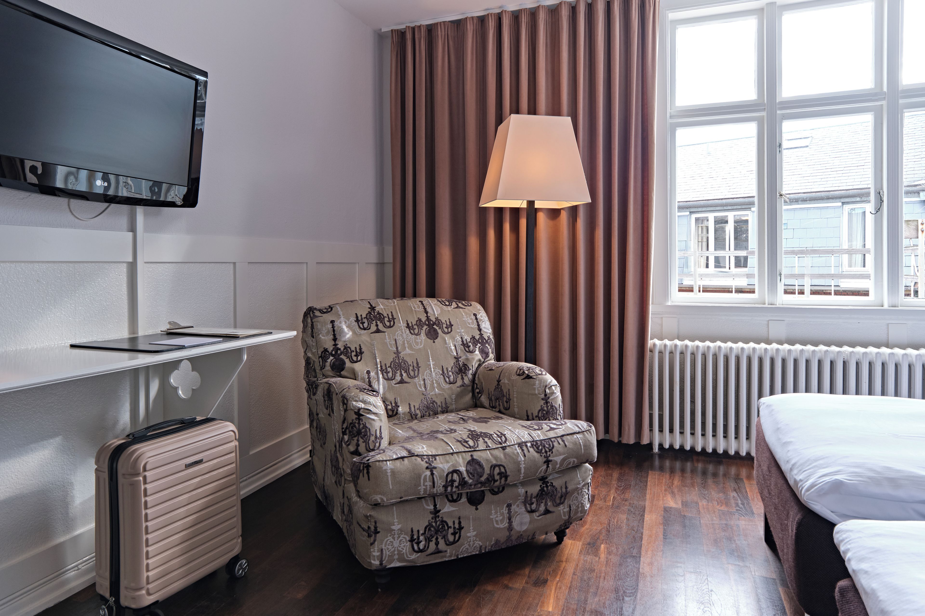 A hotel room with a bed, a desk with a flat-screen TV, a chair, a lamp, and a radiator.
