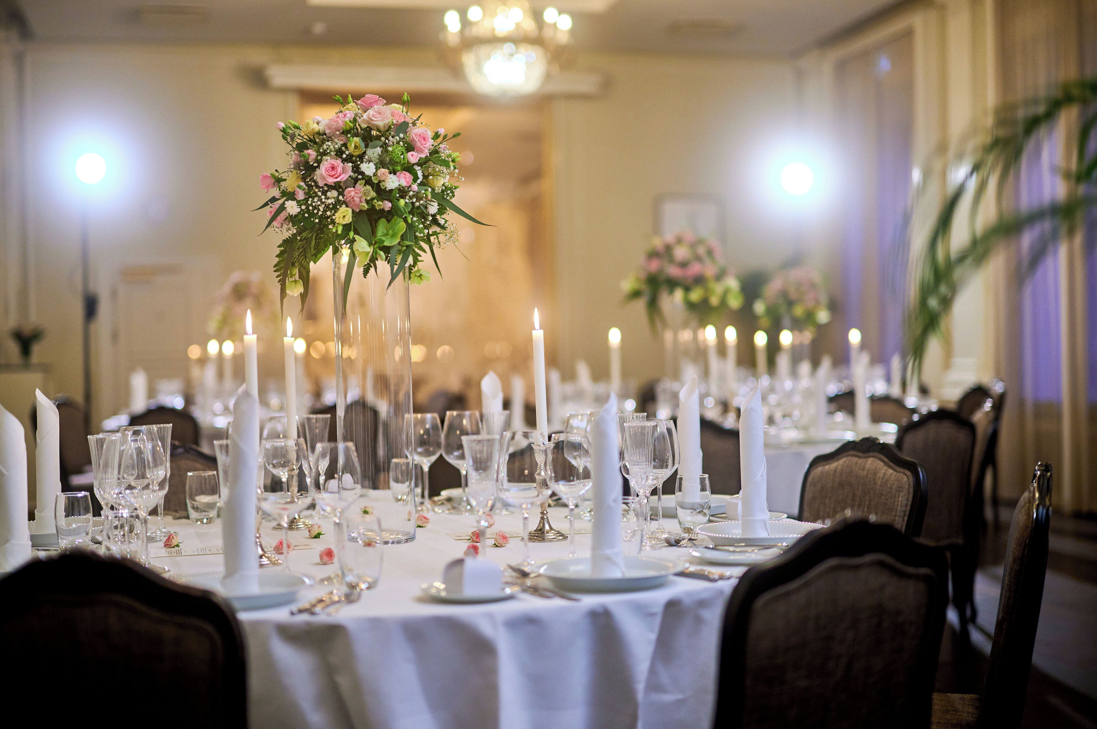 A banquet hall with tables set for a formal event, featuring tall floral centerpieces and lit candles.