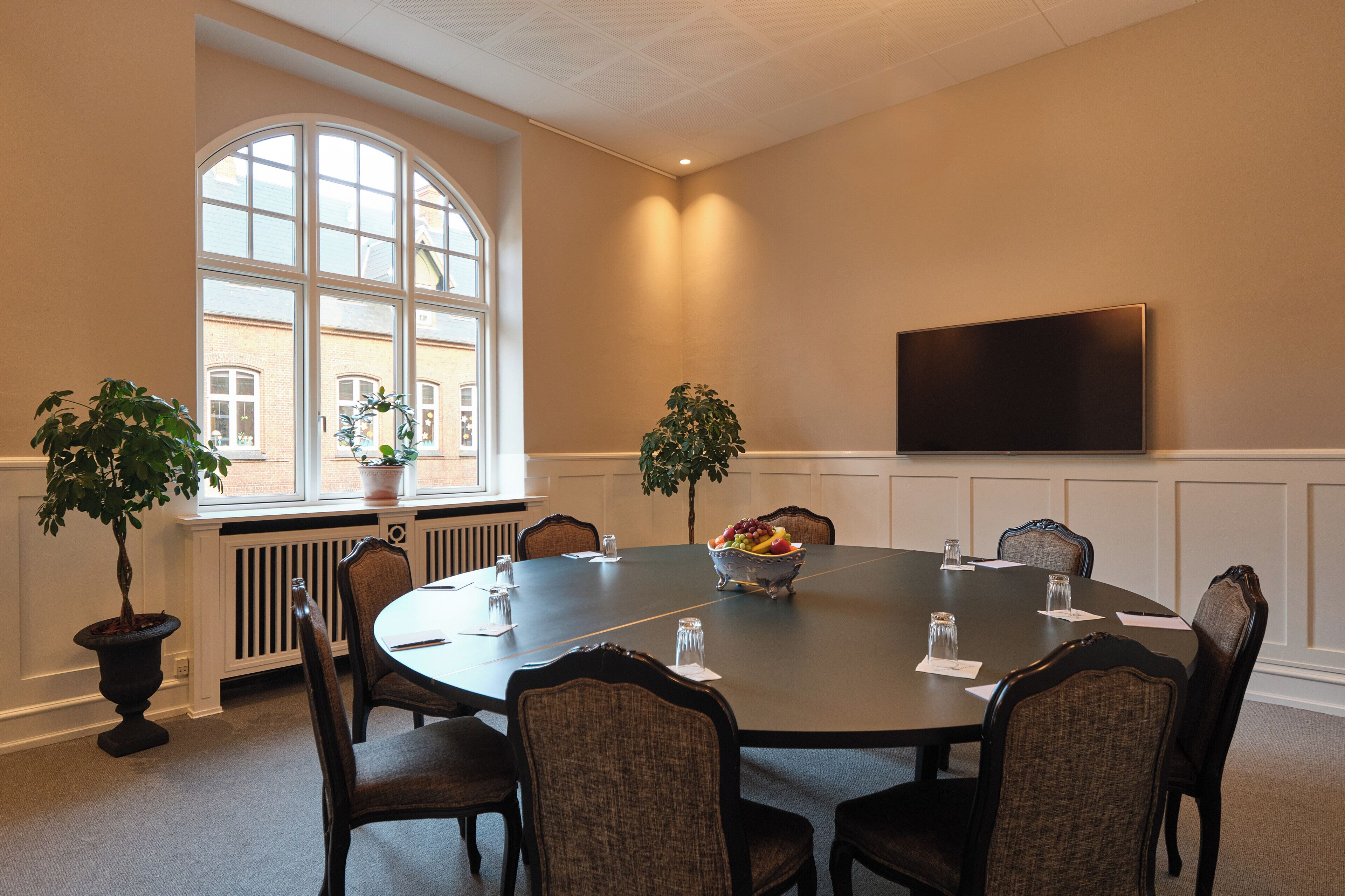 A conference room with a large table, chairs, potted plants, a fruit bowl, and a flat-screen TV on the wall.