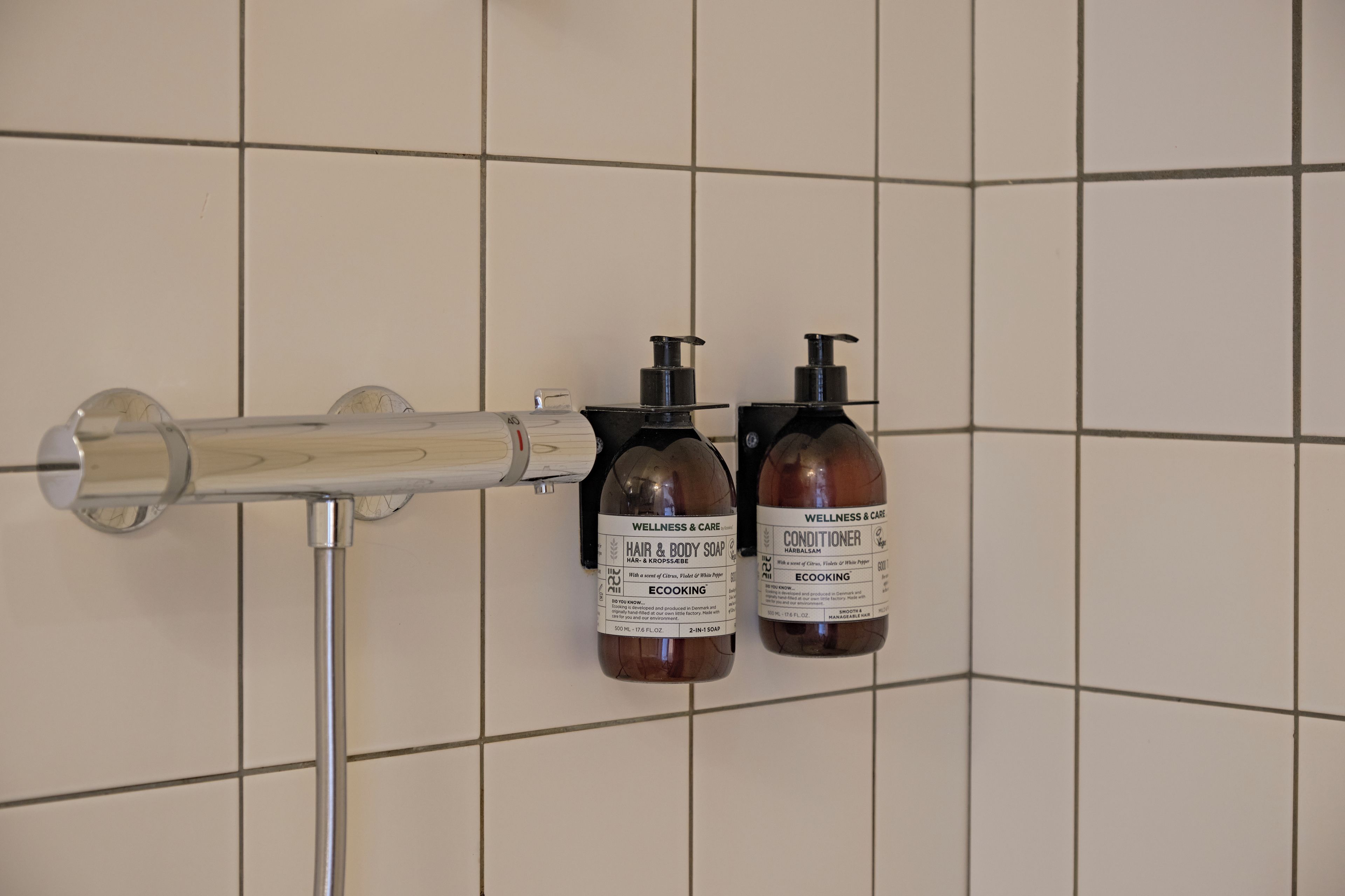 A bathroom with a shower head mounted on a wall, two bottles of hair and body soap, and a bottle of conditioner.