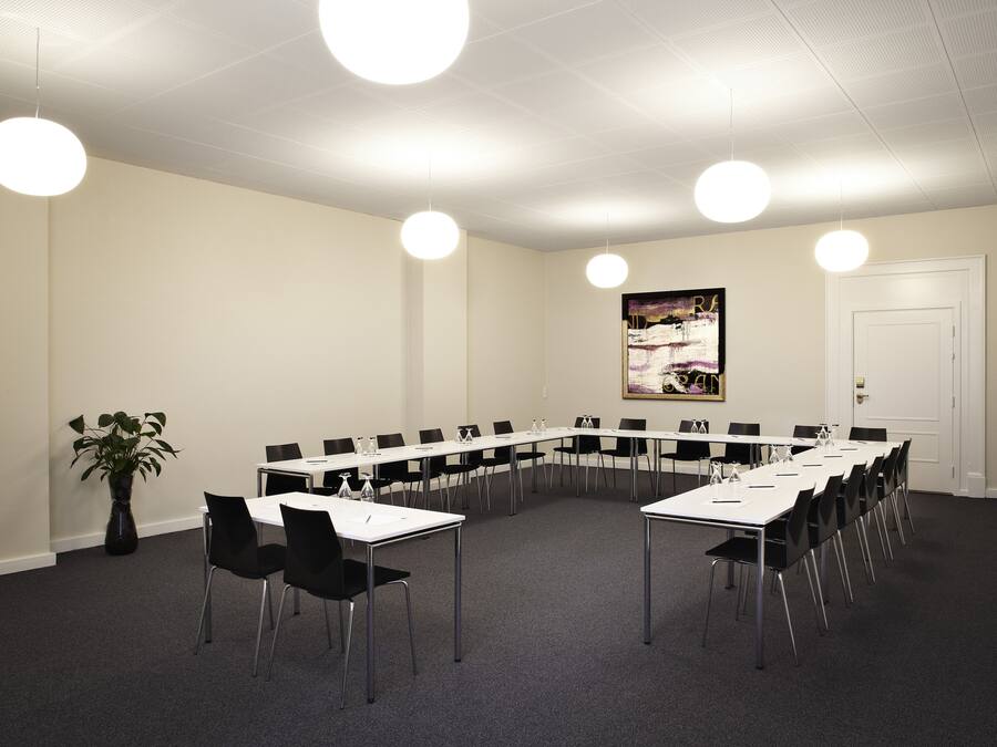 Una sala de conferencias con una larga mesa, sillas negras y mesas blancas. Hay una planta en maceta y un cuadro en la pared.