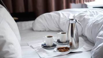 En pænt redt seng med hvide sengetøj, en sølvfarvet kaffepotte, to hvide kaffekopper og en tallerken med småkager.