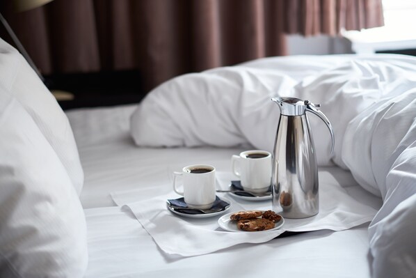 A neatly made bed with white linens, a silver coffee pot, two white cups of coffee, and a plate with cookies.