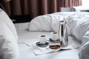 En pænt redt seng med hvide sengetøj, en sølvfarvet kaffepotte, to hvide kaffekopper og en tallerken med småkager.