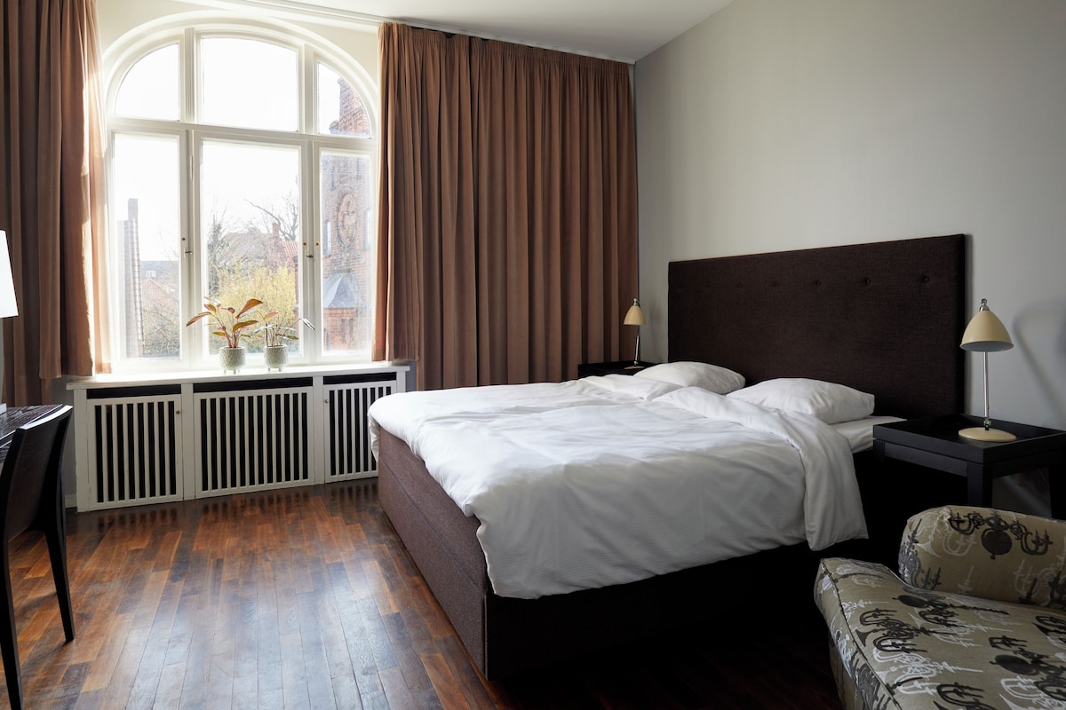 Un dormitorio con un ventanal grande, una cama, una mesita de noche con una lámpara, una silla y un radiador.