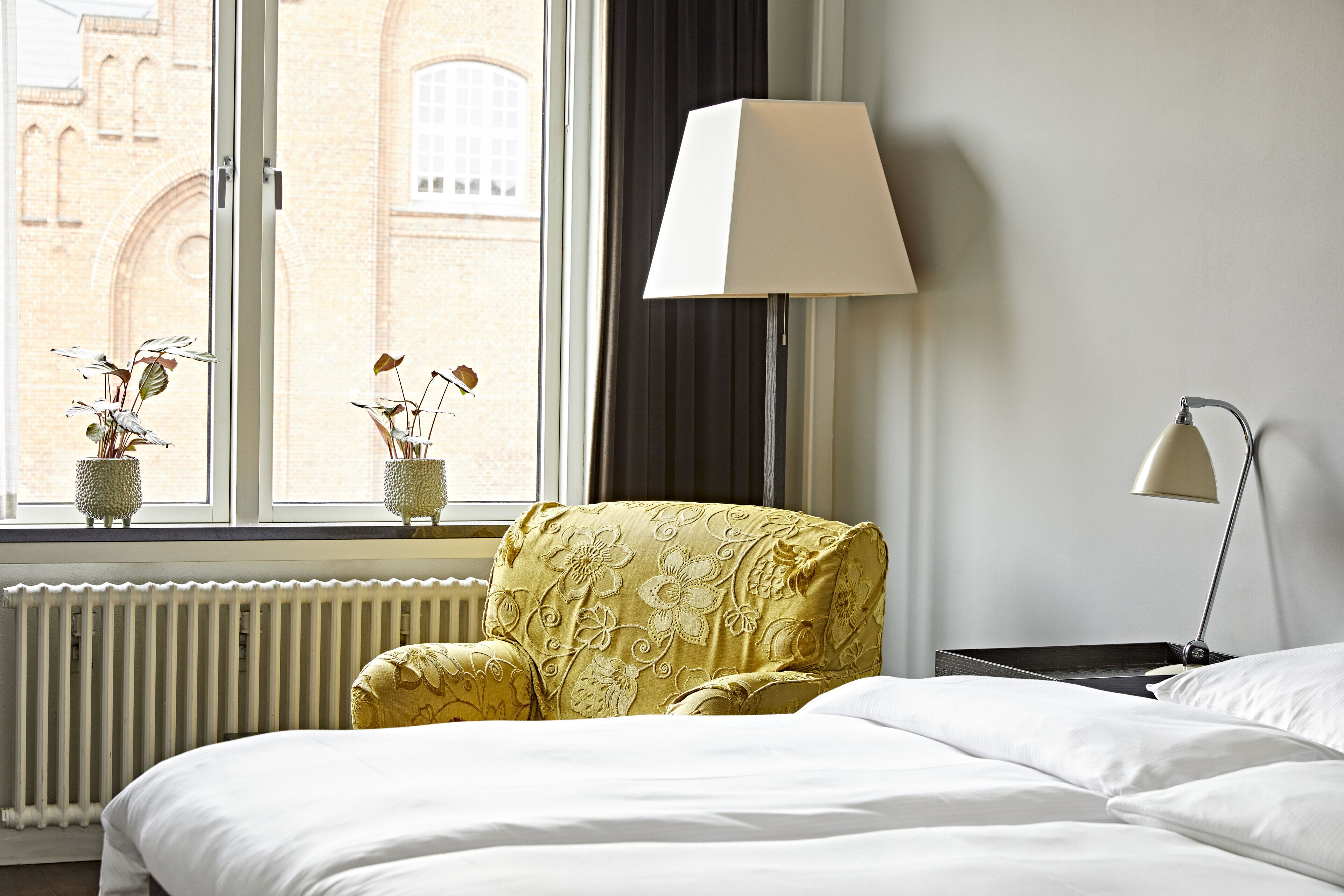 Camera d'albergo con un letto, una sedia, una lampada e una finestra vista su un edificio in mattoni.