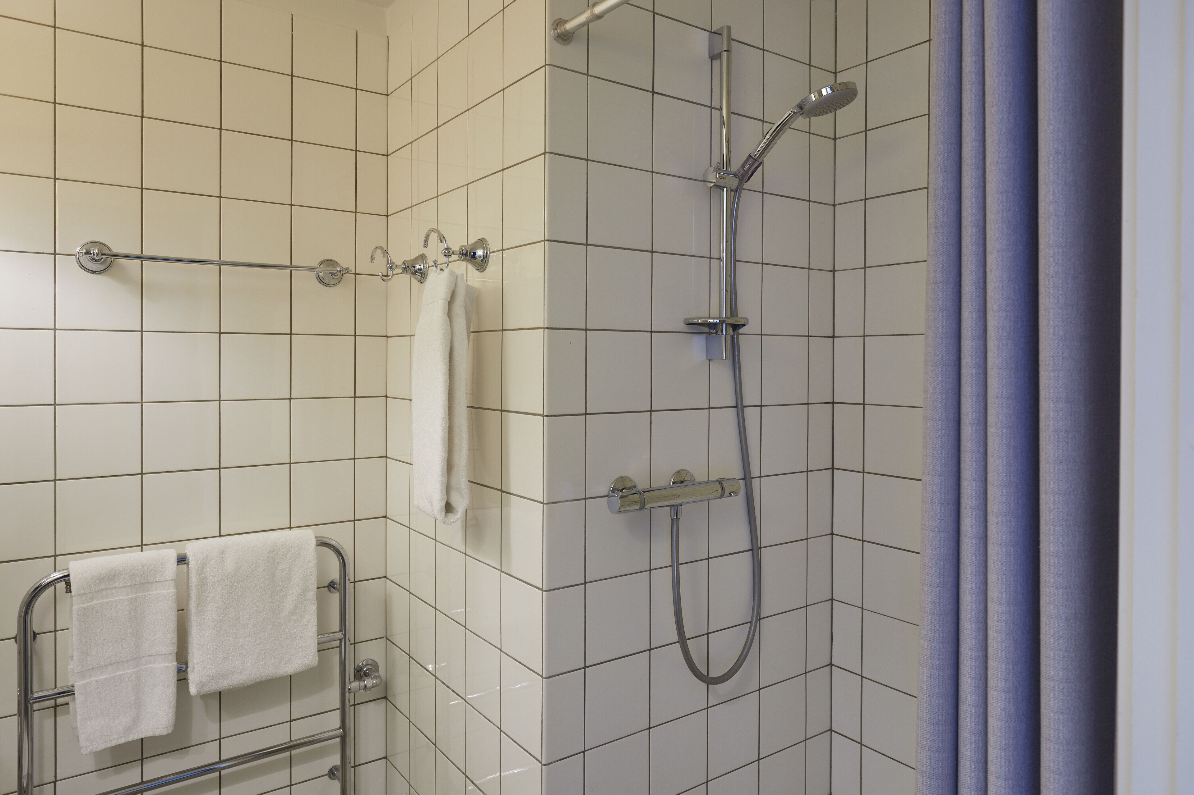 A bathroom with white tiled walls, a shower with a handheld showerhead, and a towel rack with two towels.
