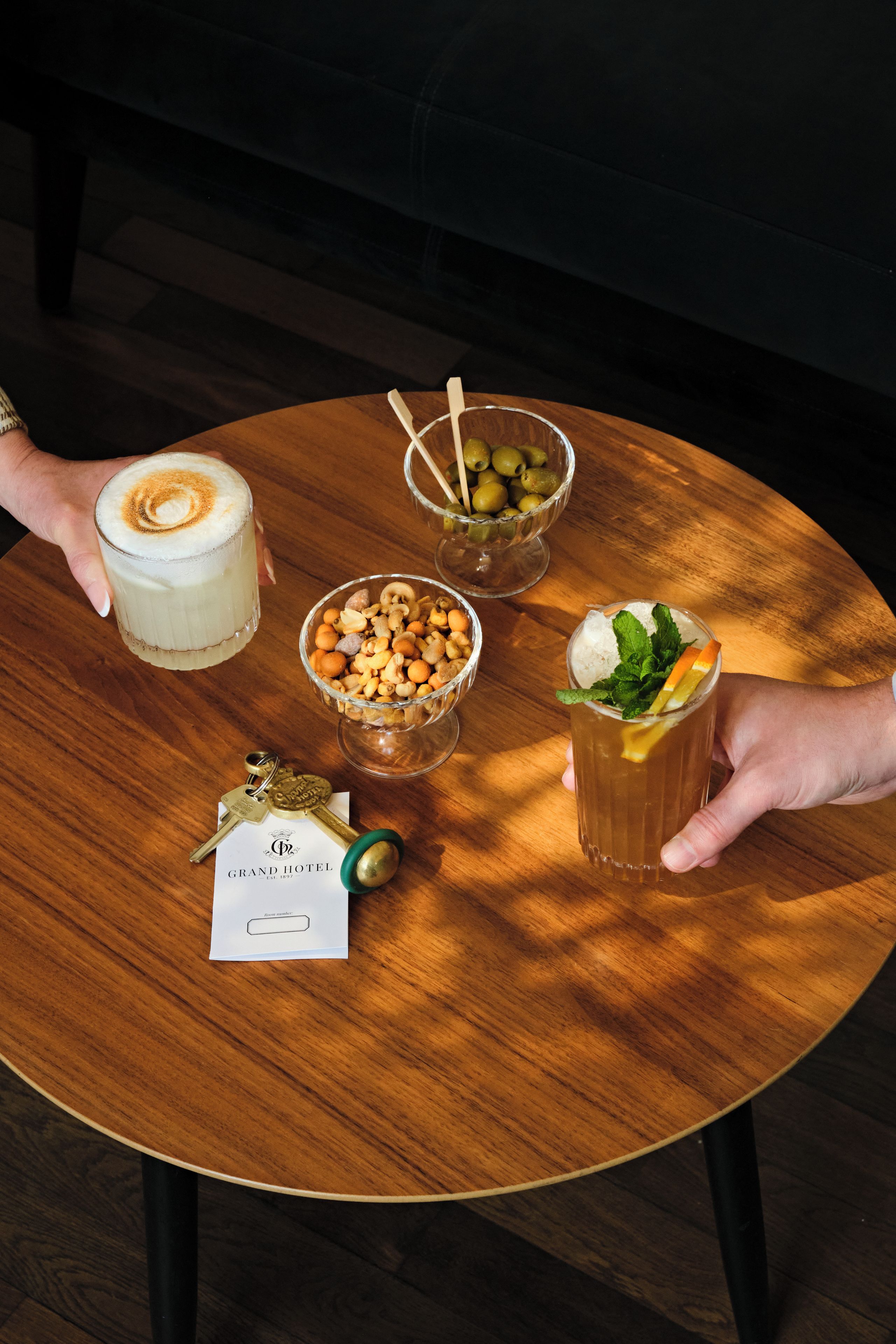 A round wooden table with four items: a glass of coffee with latte art, a bowl of mixed nuts, a glass of iced tea with lemon and mint, and a brass keychain with a hotel key.