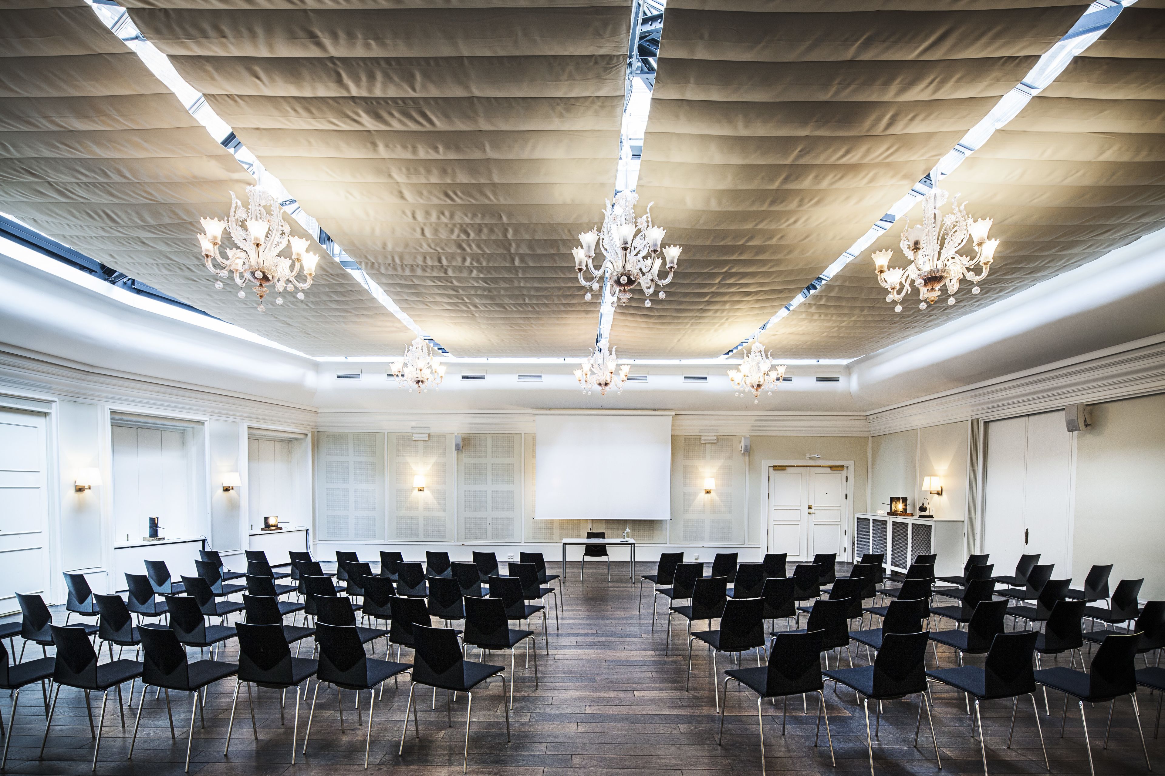 A conference room with rows of black chairs, a large screen, and chandeliers.