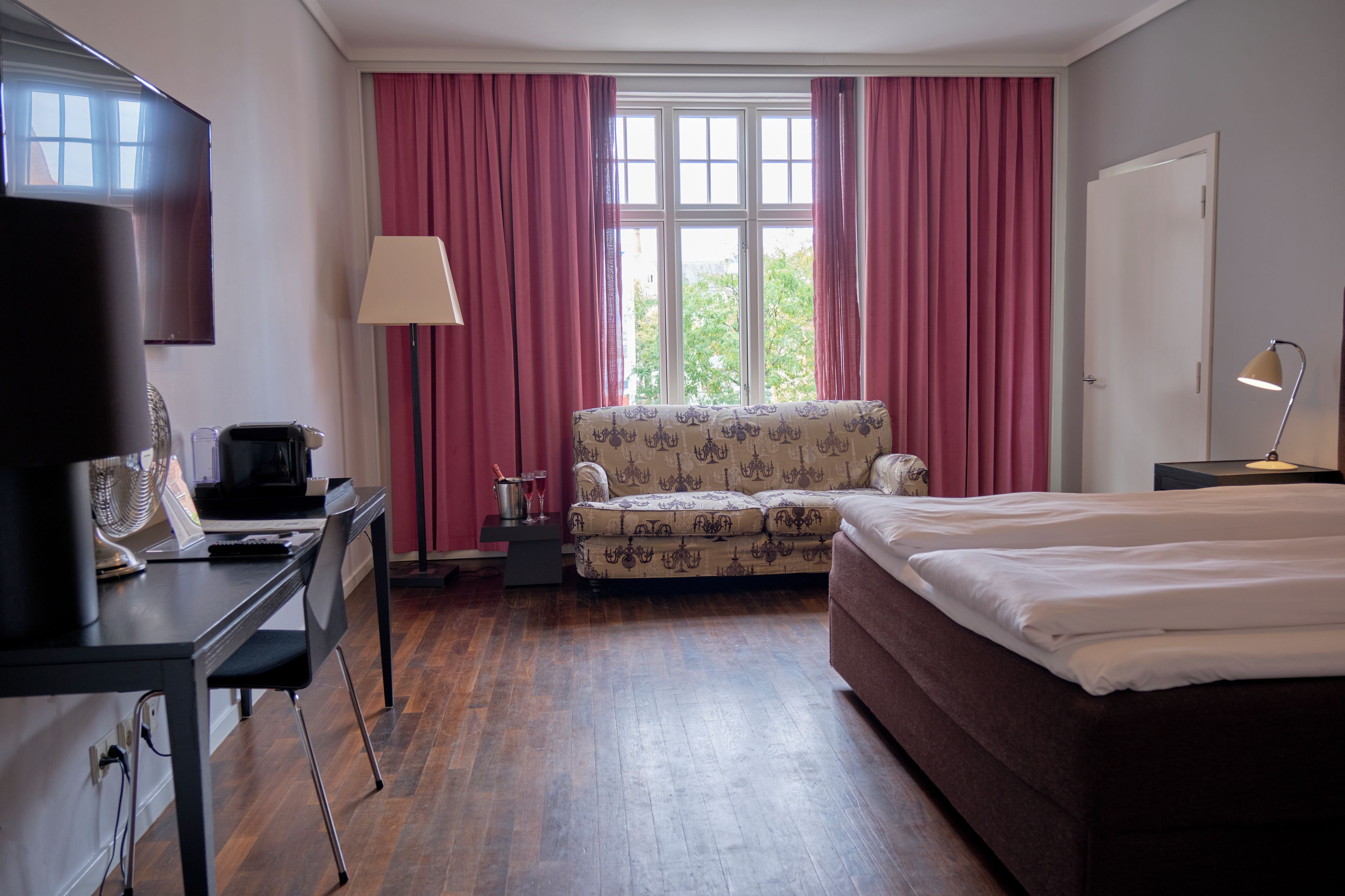 Una habitación de hotel con una cama, un escritorio con computadora, una silla, una lámpara y un ventanal con cortinas.