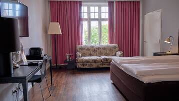 Una habitación de hotel con una cama, un escritorio con computadora, una silla, una lámpara y un ventanal con cortinas.