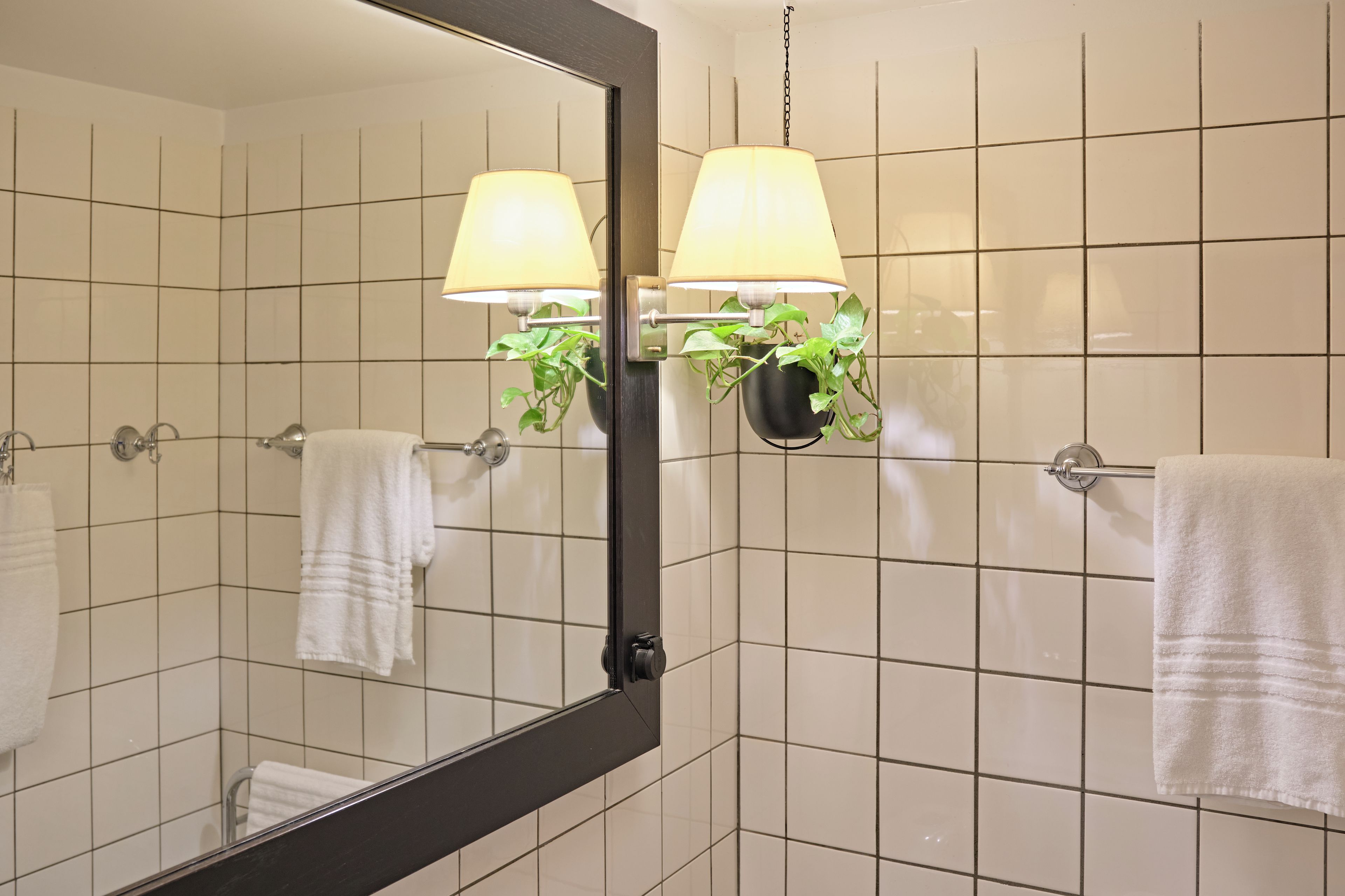 Un baño con paredes de azulejos blancos, un espejo grande, dos lámparas empotradas y dos toallas colgadas.