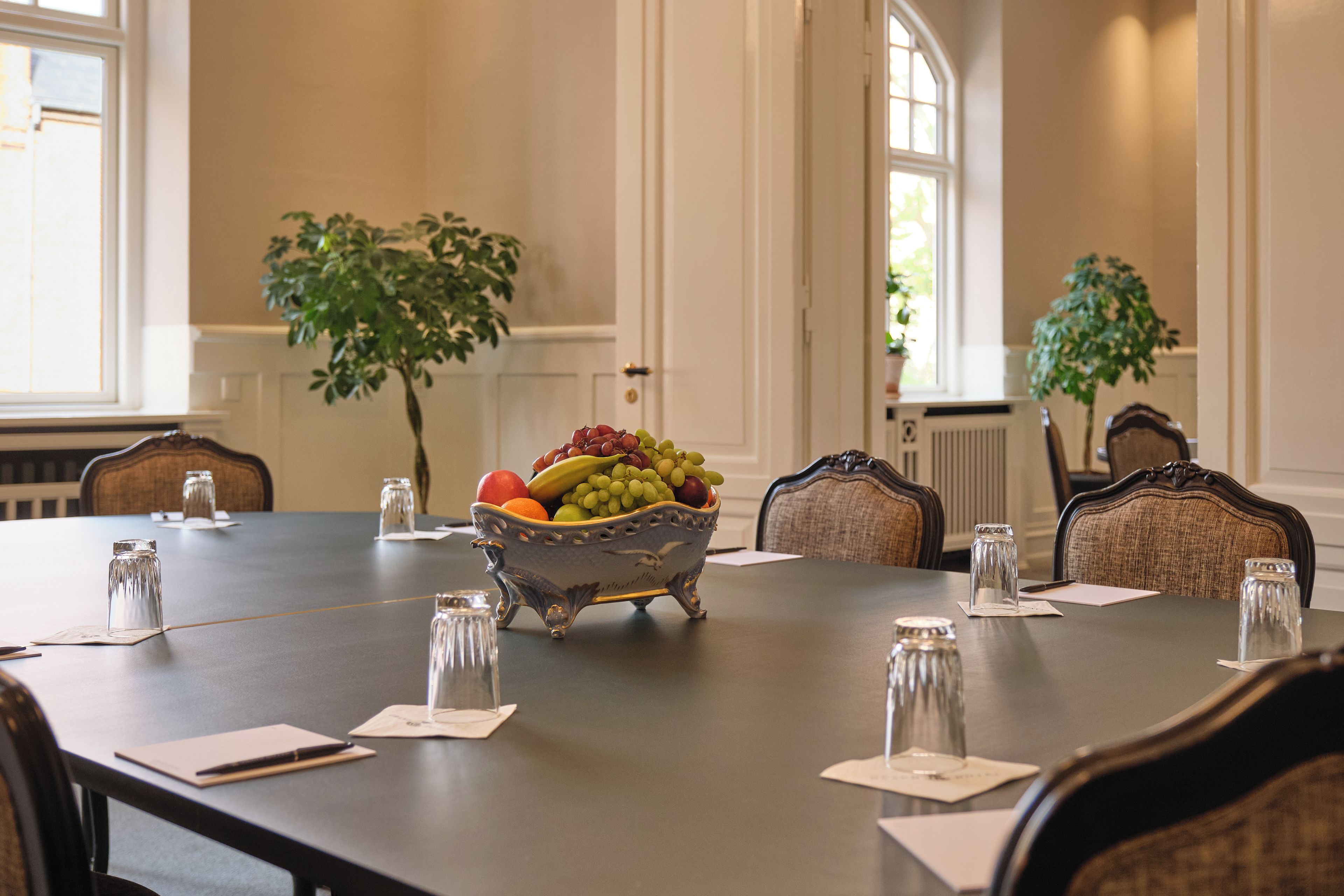 A conference table with a fruit bowl, glasses, and notepads in a room with chairs and windows.