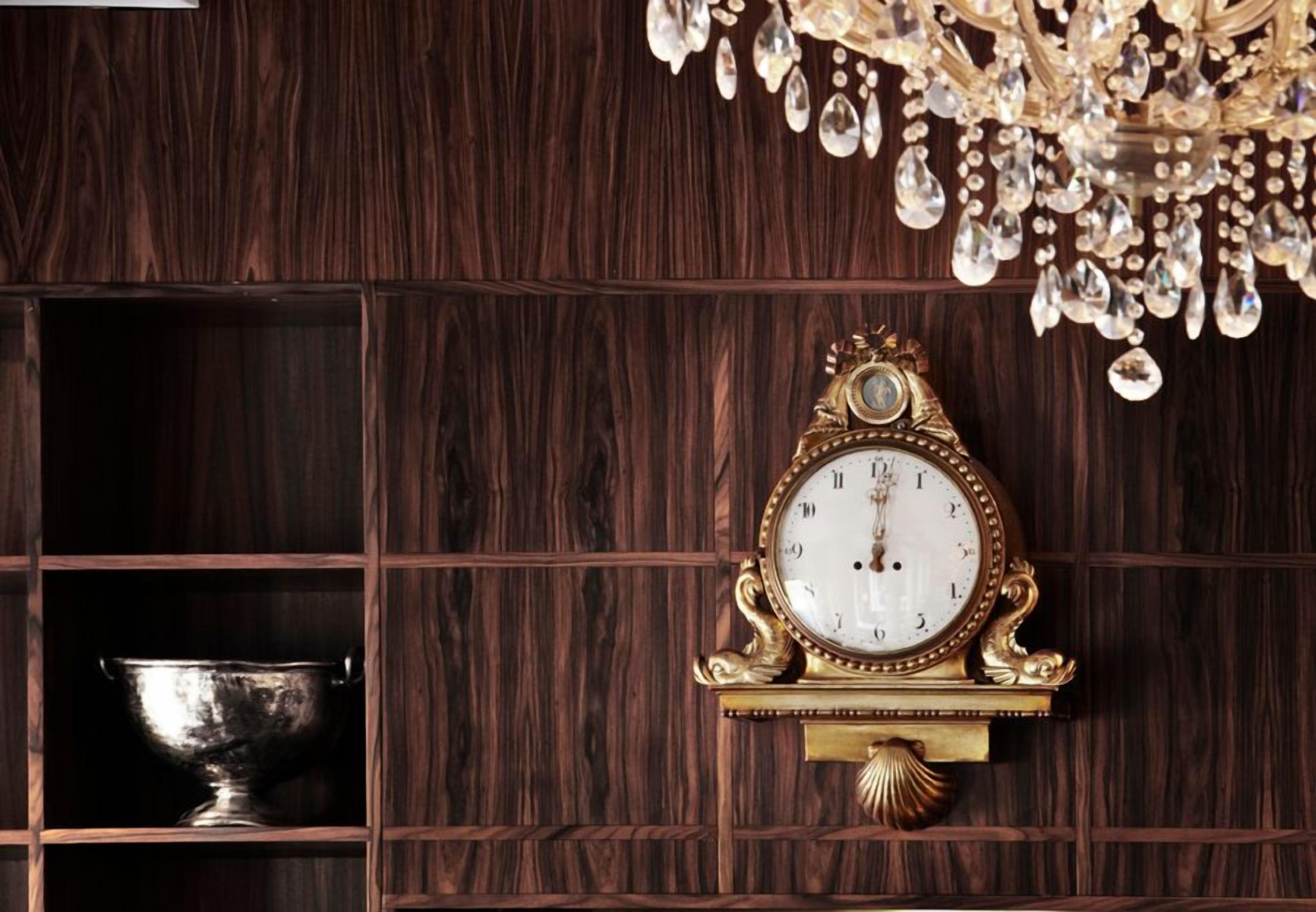 Un reloj de pared con base decorativa, un cuenco de plata en un aparador de madera y una araña con detalles de cristal.