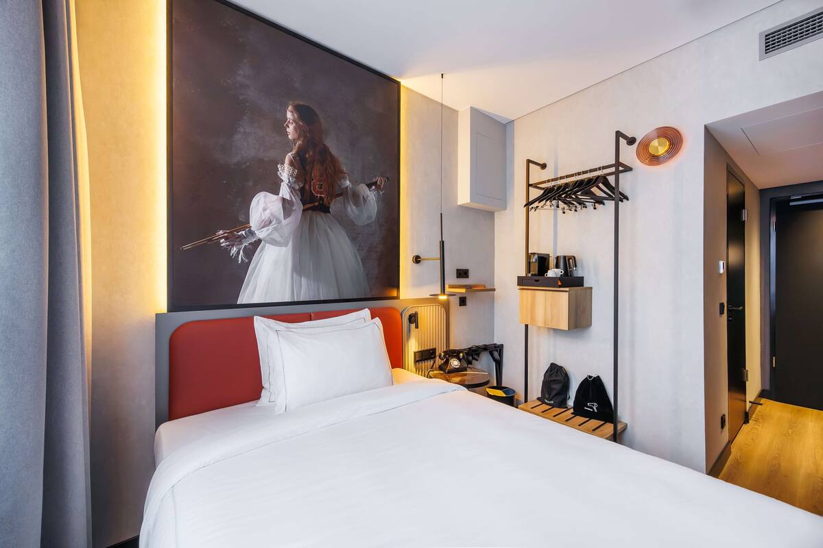 Habitación de hotel con una cama grande, cabecera roja, un cuadro en la pared, un perchero y piso de madera.