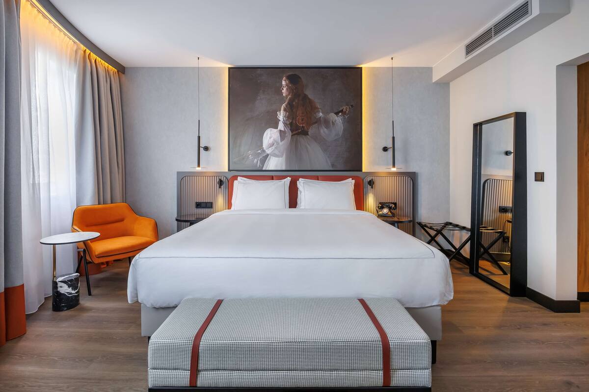 Una habitación de hotel moderna con una cama grande, una mesita pequeña, una silla naranja y un cuadro grande en la pared.