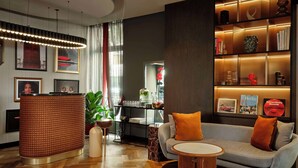 A modern hotel lobby with a reception desk, a sofa with orange cushions, a bookshelf with books and decorative items, and a large window with curtains.