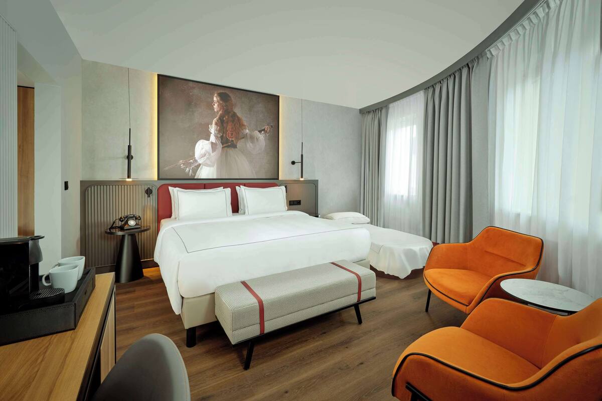 Habitación de hotel con una cama grande, una mesita de noche con lámpara, una pequeña mesa con silla y un ventanal con cortinas.