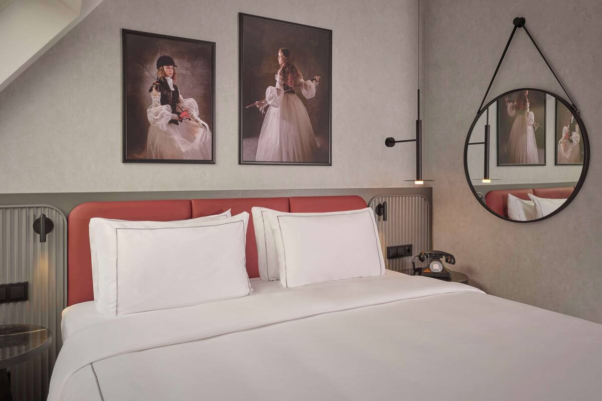 Habitación de hotel con una cama grande, cabecera roja, dos retratos enmarcados en la pared, un espejo redondo y una mesita de noche con teléfono.
