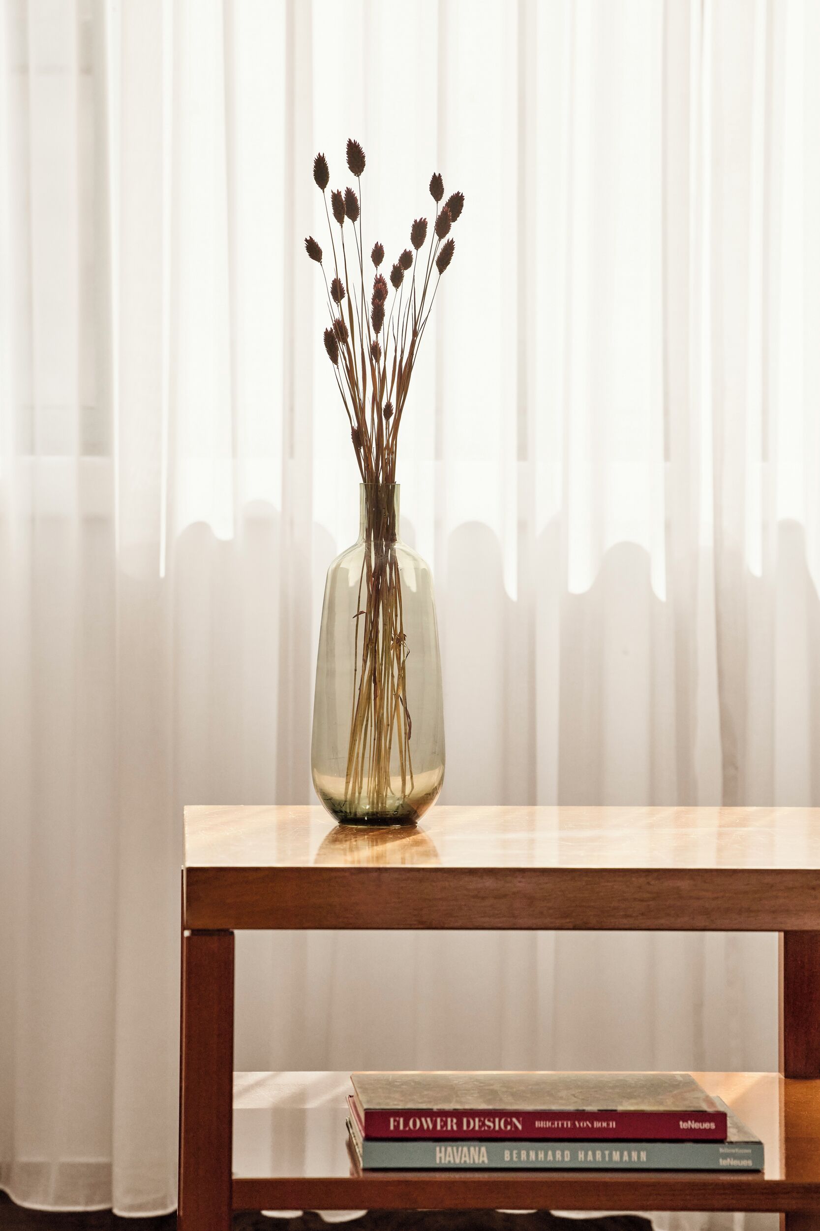 A clear view of a wooden table with a vase containing dried flowers and two books titled "Flower Design" and "Havana" by Bernhard Hartmann.