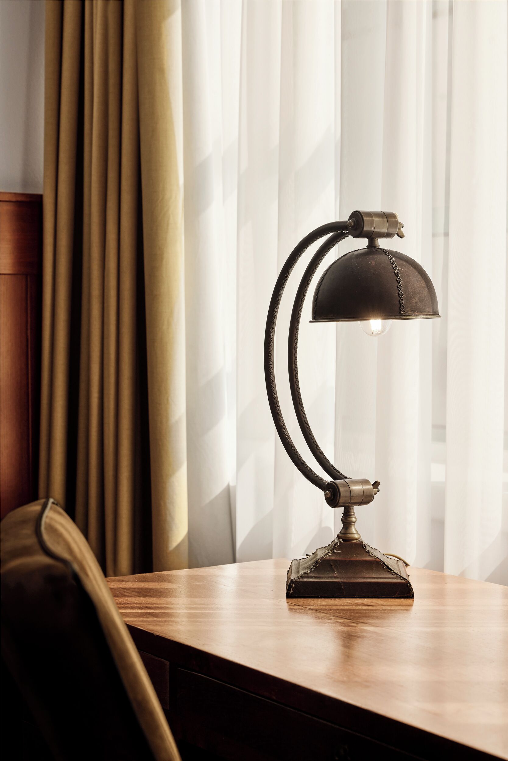 Eine Schreibtischlampe mit gebogenem Arm und Lampenschirm auf einem Holztisch in einem Zimmer mit Fenster und Vorhängen.