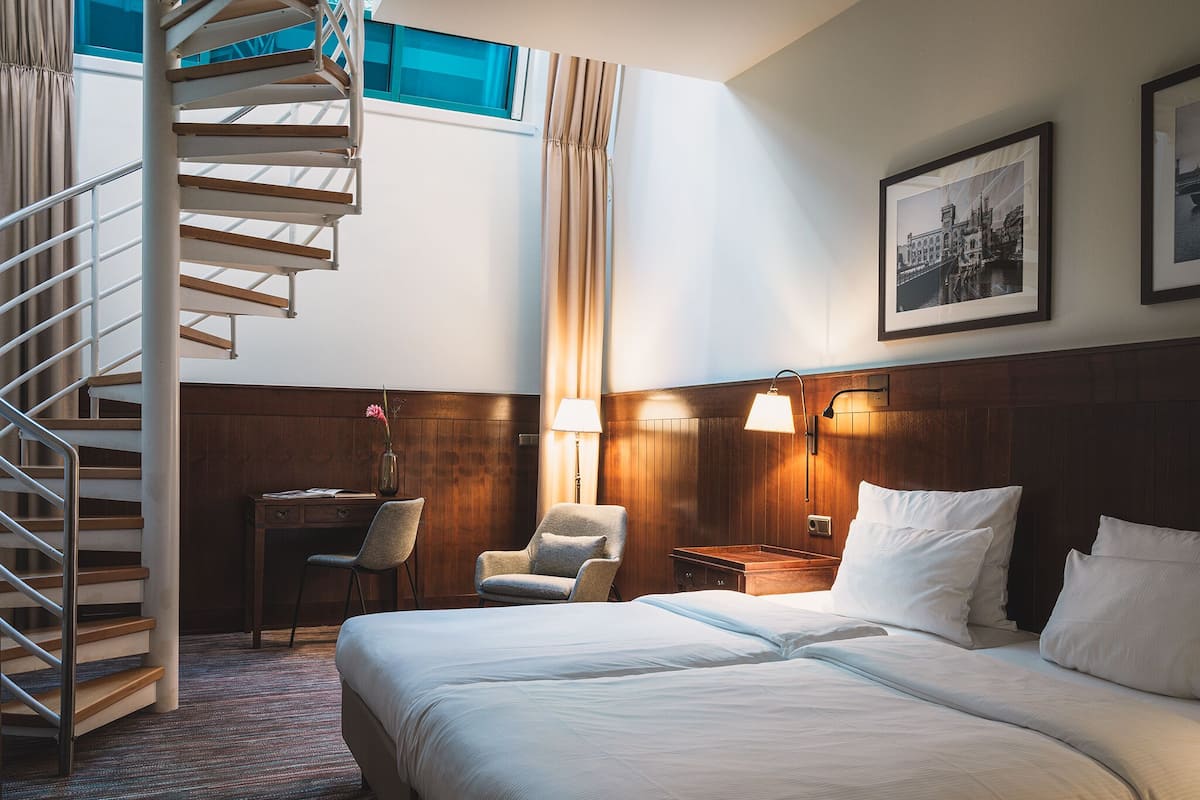 Una habitación de hotel con una cama, un escritorio, una silla, una lámpara y una escalera.