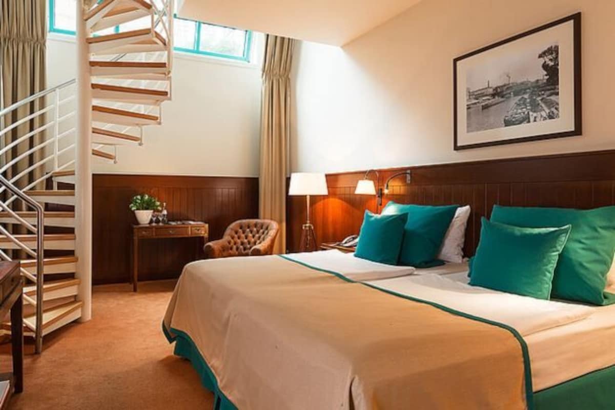 A hotel room with a large bed, a wooden headboard, a desk with a lamp, a chair, a small table with a plant, and a spiral staircase.