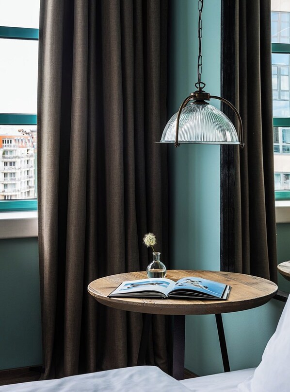 Una habitación de hotel con una mesa redonda de madera, un libro abierto, un jarrón con una flor y una lámpara colgante.