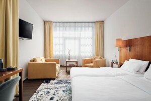 A hotel room with a bed, two armchairs, a TV, a small table with a lamp, and a window with curtains.