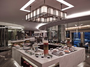 Eine moderne Hotelhalle mit einer zentralen Bar, die verschiedene Speisen und Getränke anbietet, einem großen Kronleuchter und einer sitzbaren Glasgalerie.