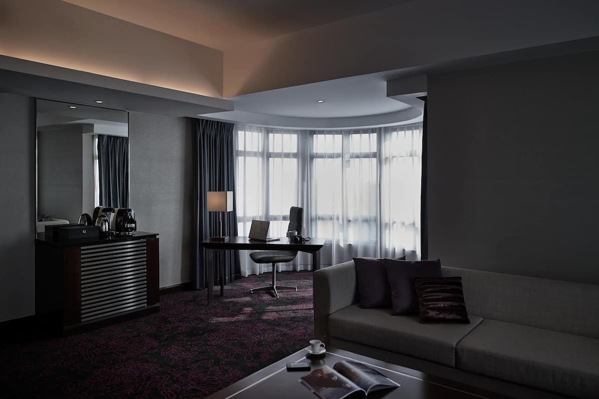 Moderni hotellihuone, jossa on tumma matto, sohva, työpöytä kannettavalla tietokoneella, kahvinkeitin ja lamppu.