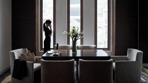 Una habitación de hotel con un ventanal grande, una mesa de comedor con sillas, un florero con flores y un bolso sobre una silla.